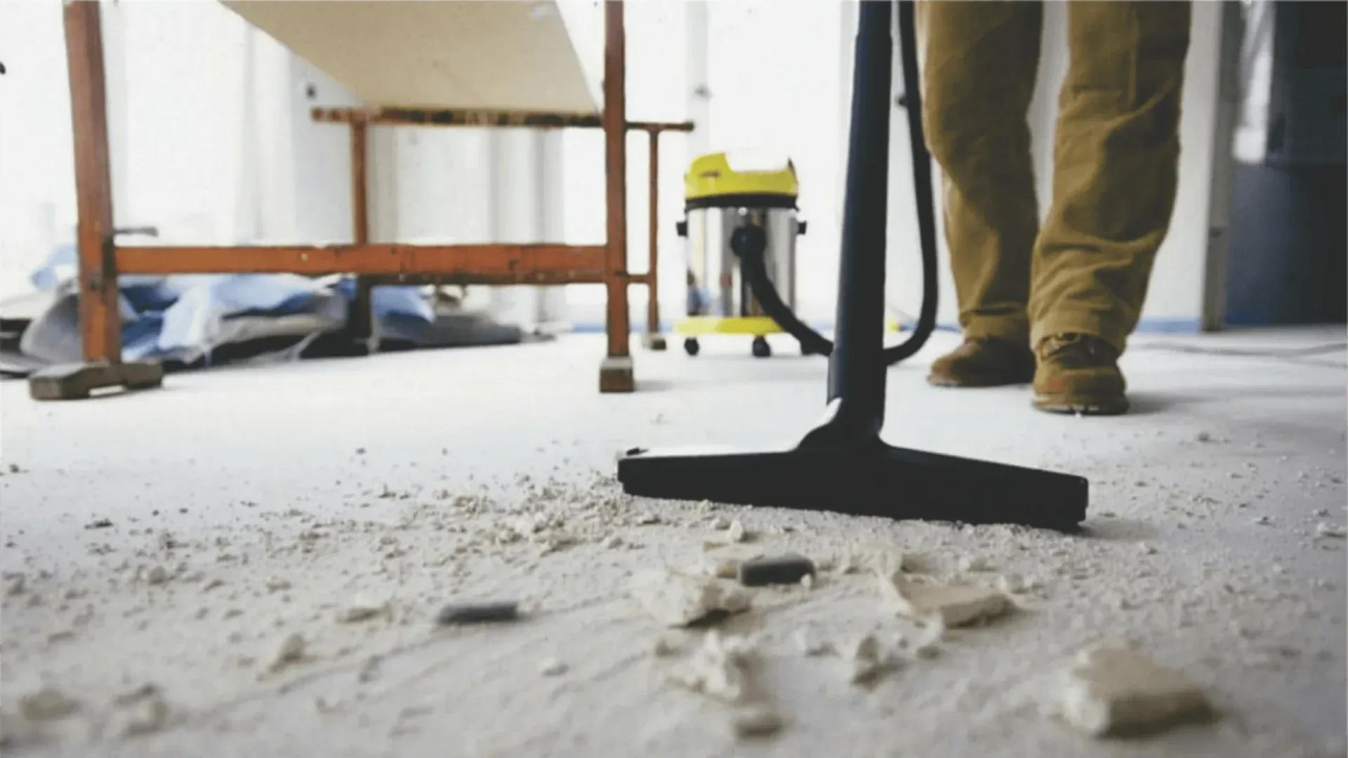 Person working on a post-construction clean, vacuuming a dusty floor with a yellow shop vacuum in a construction zone.