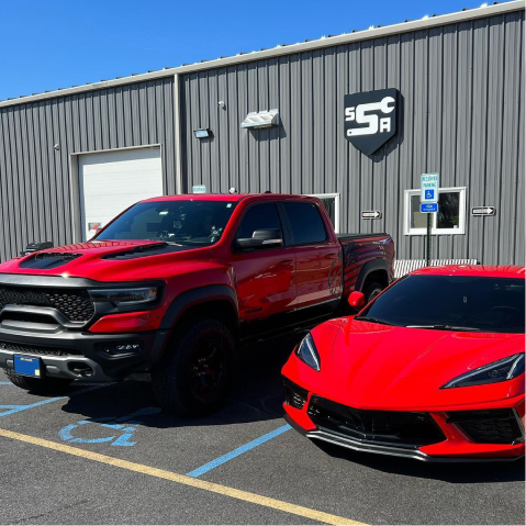 Red pickup and red sports car parked outside a gray automotive shop.