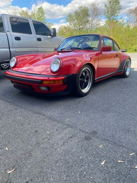 Red classic Porsche 911 parked on asphalt beside a silver truck, with trees in the background.