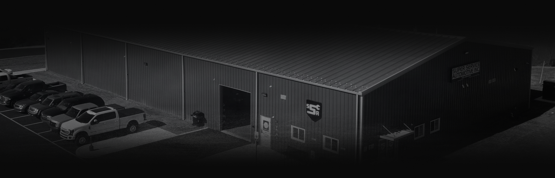Dark industrial warehouse exterior with parked cars along the left side and loading doors on the right