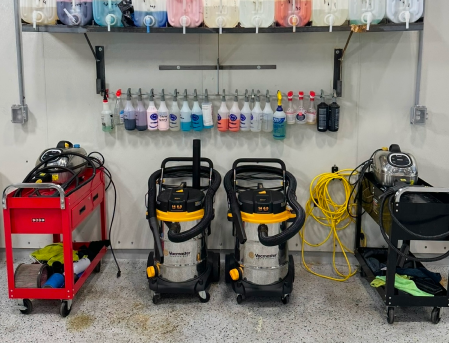 Cleaning supply room with two wet/dry vacuums, red cart, hoses, and bottles on wall shelves.