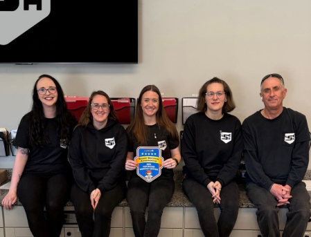 Five people in black sweatshirts sit on a bench; the center person holds a blue-and-yellow award plaque.