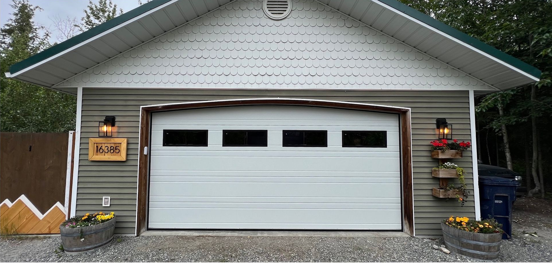 Garage with a white door, two wall lamps, and flower boxes. Brown wooden mountain fence.
