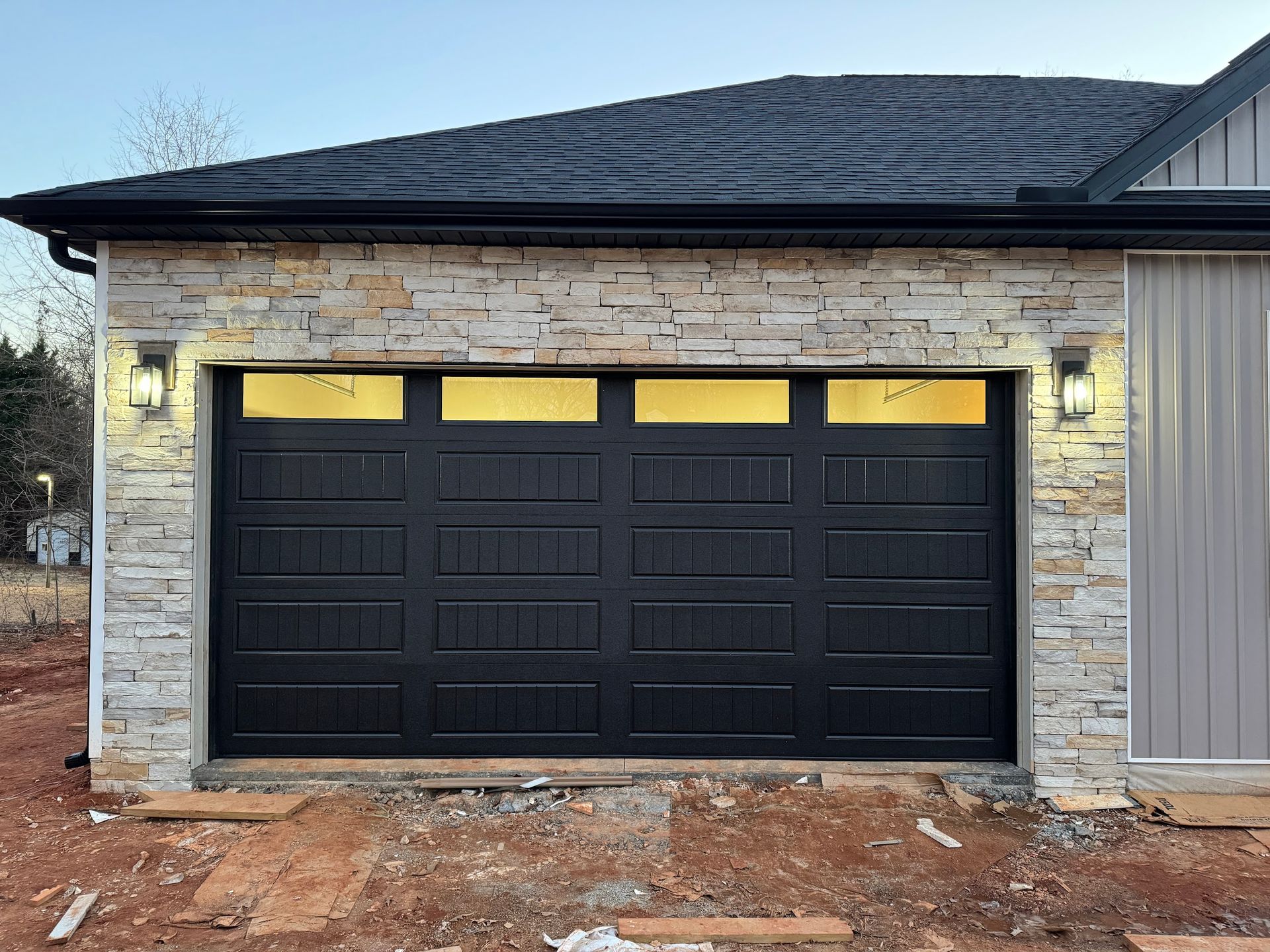 Black garage door with stone facade, lit by outdoor wall lamps.