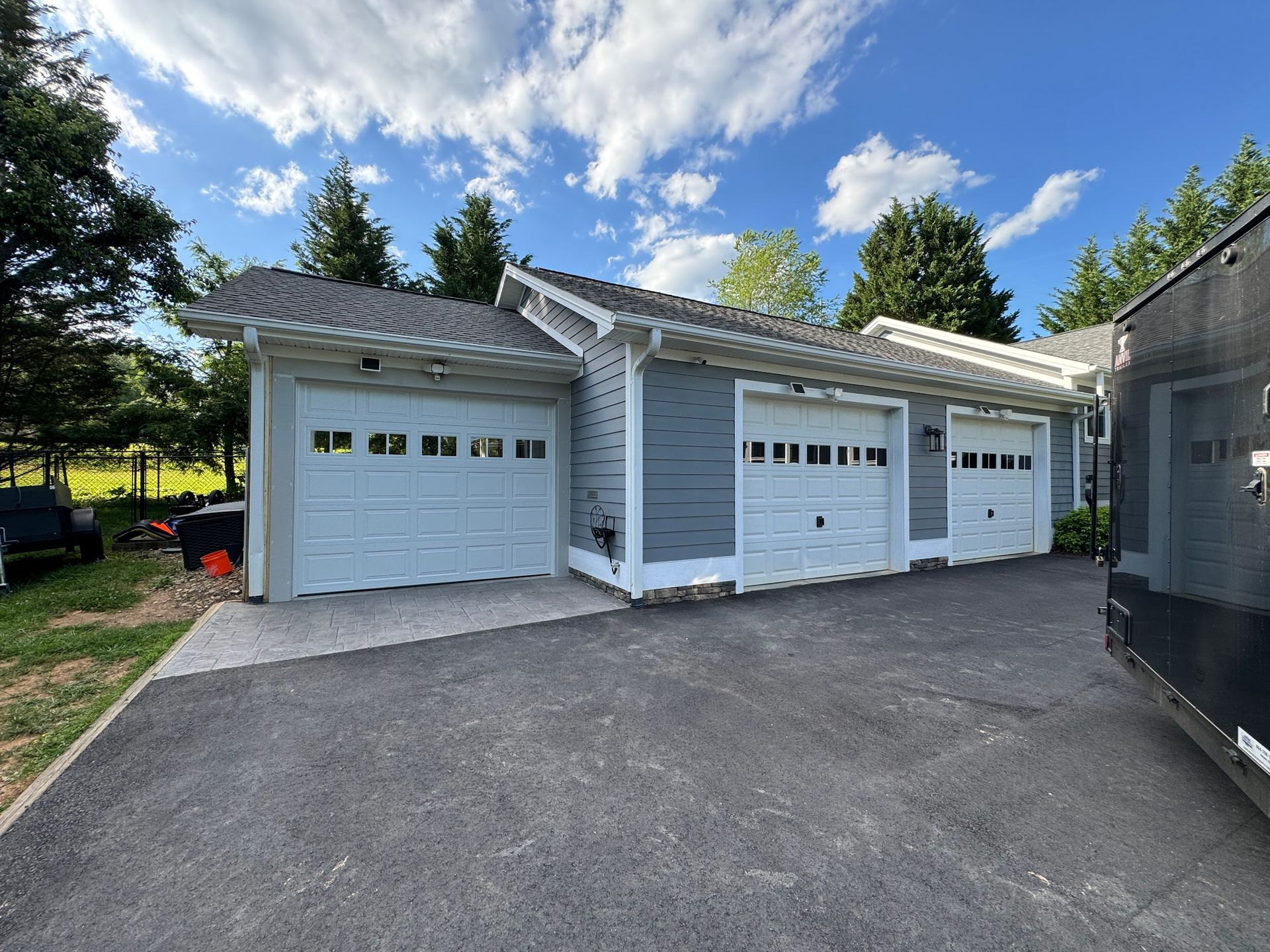 Three-car garage with white doors and gray siding. Asphalt driveway, blue sky, and green trees.