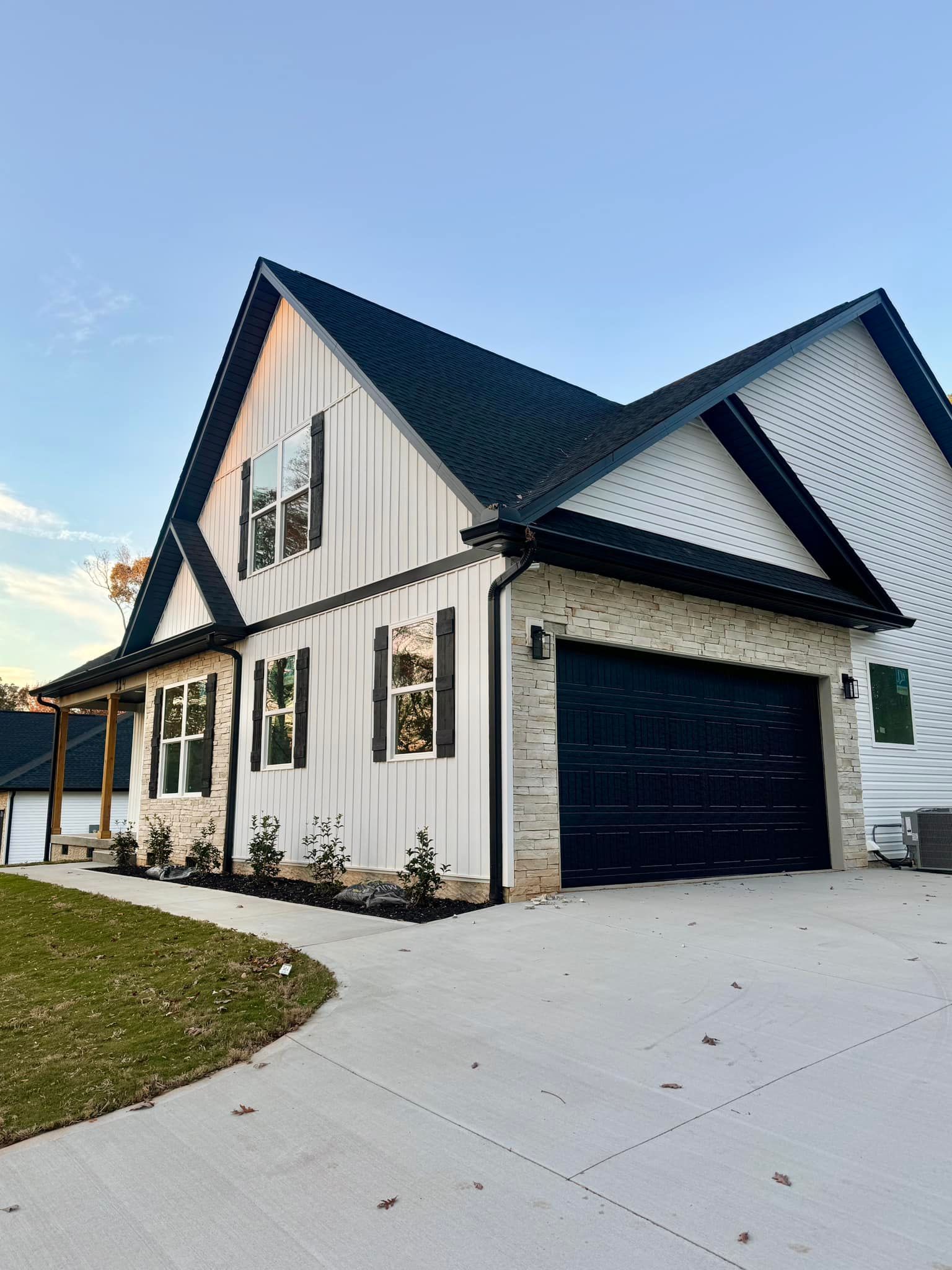 White farmhouse with black roof and garage door; brick accents; concrete driveway.