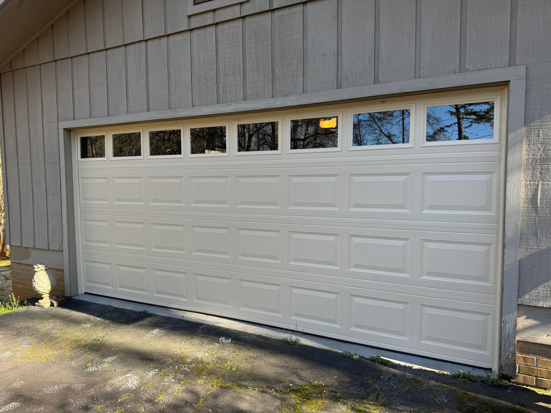 Beige garage door with windows, set in a building with grey siding and surrounded by trees.