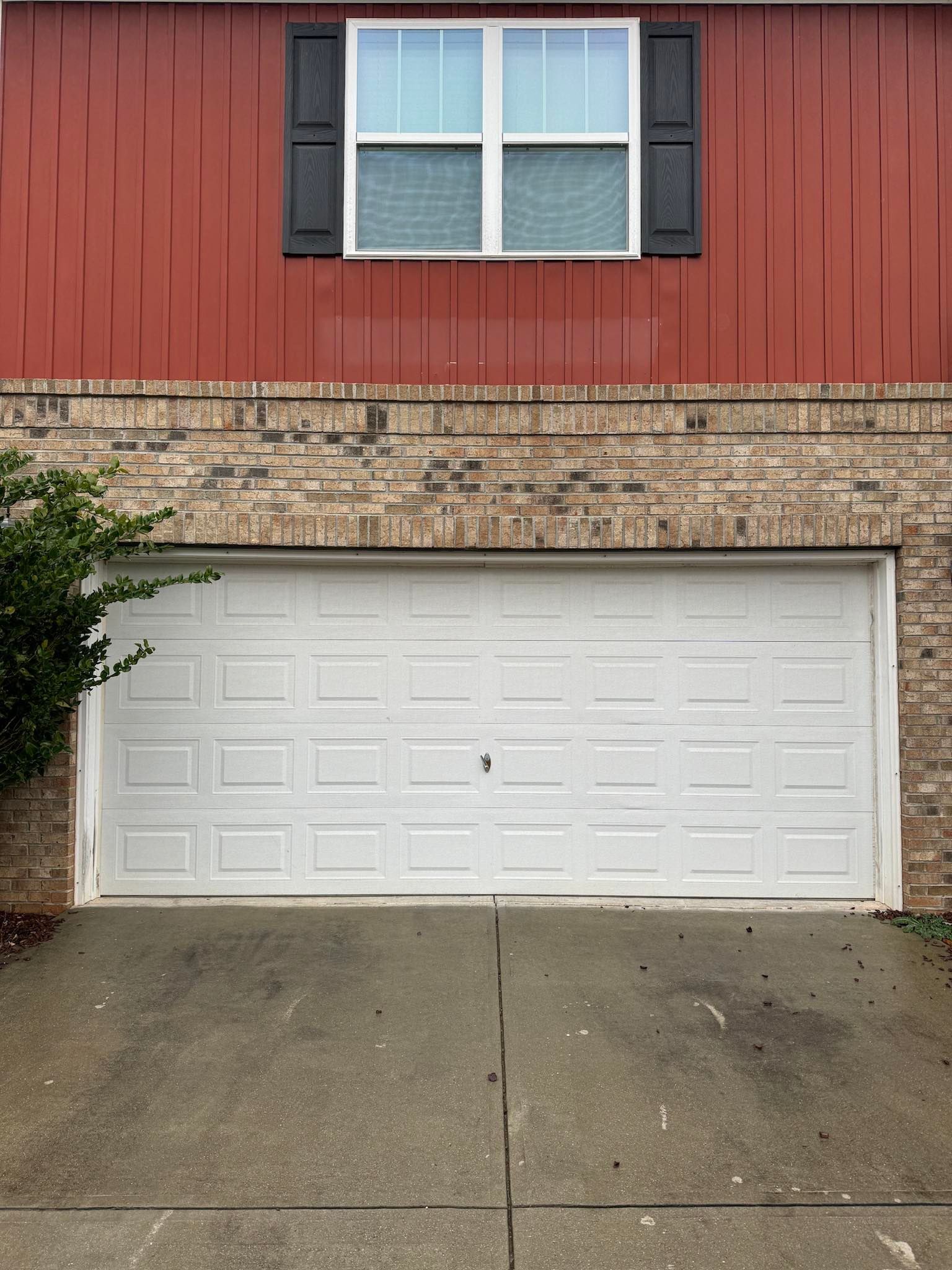 White garage door on a brick and red-paneled house, with a window and black shutters above.