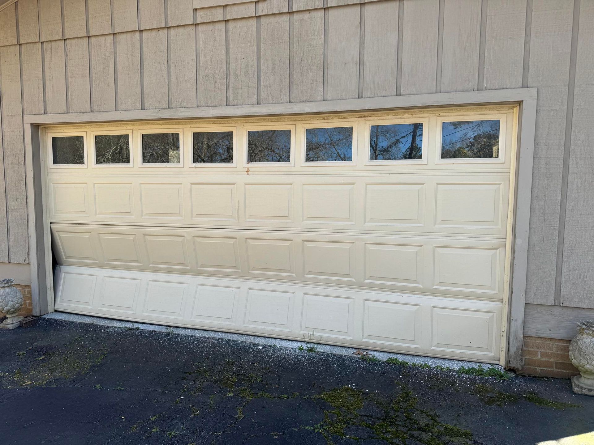 Garage door, partially open, cream-colored panels, slight bend at bottom left. Building has beige siding.