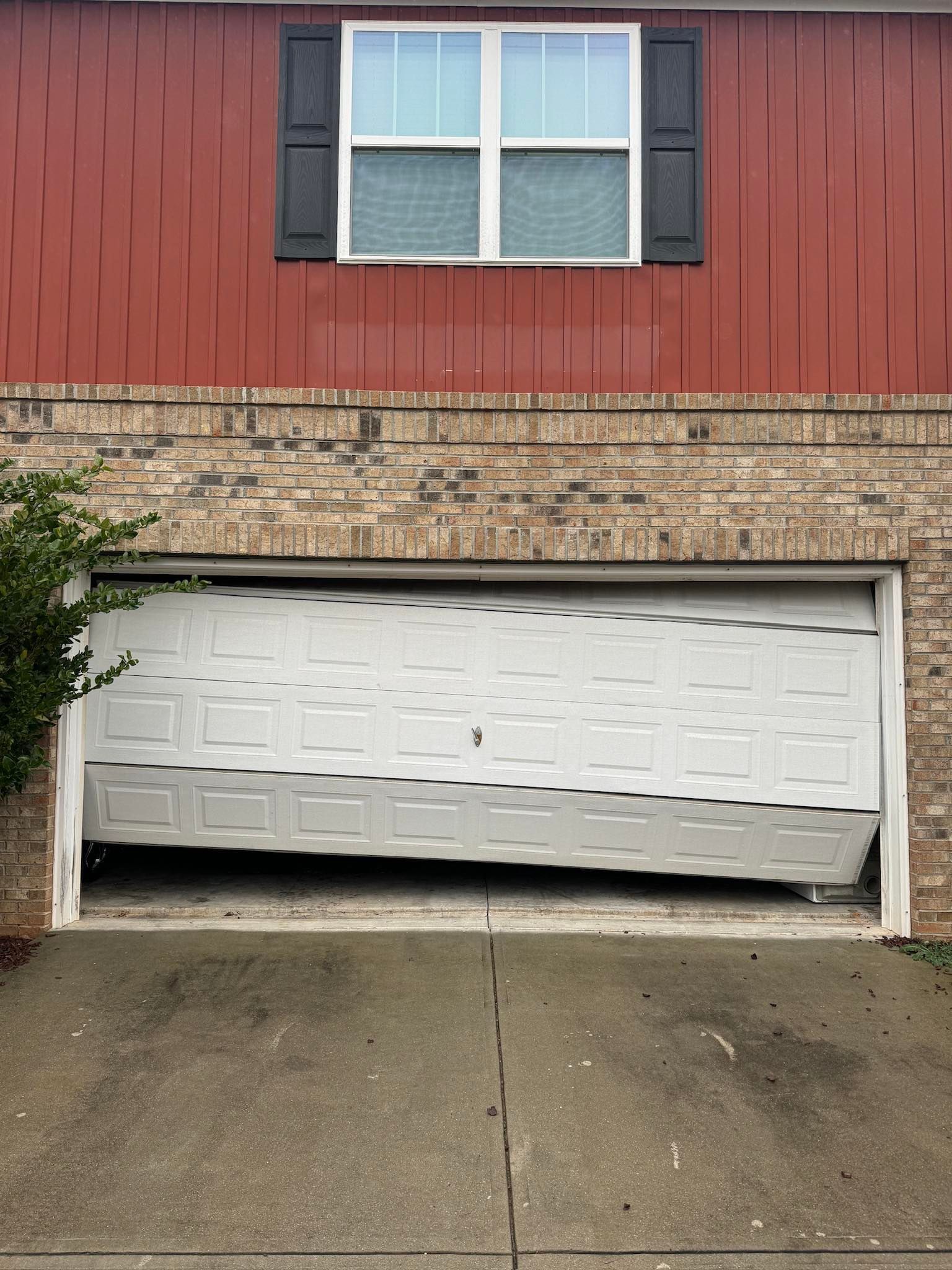 Garage door partially open and crumpled. Brick and red siding house.