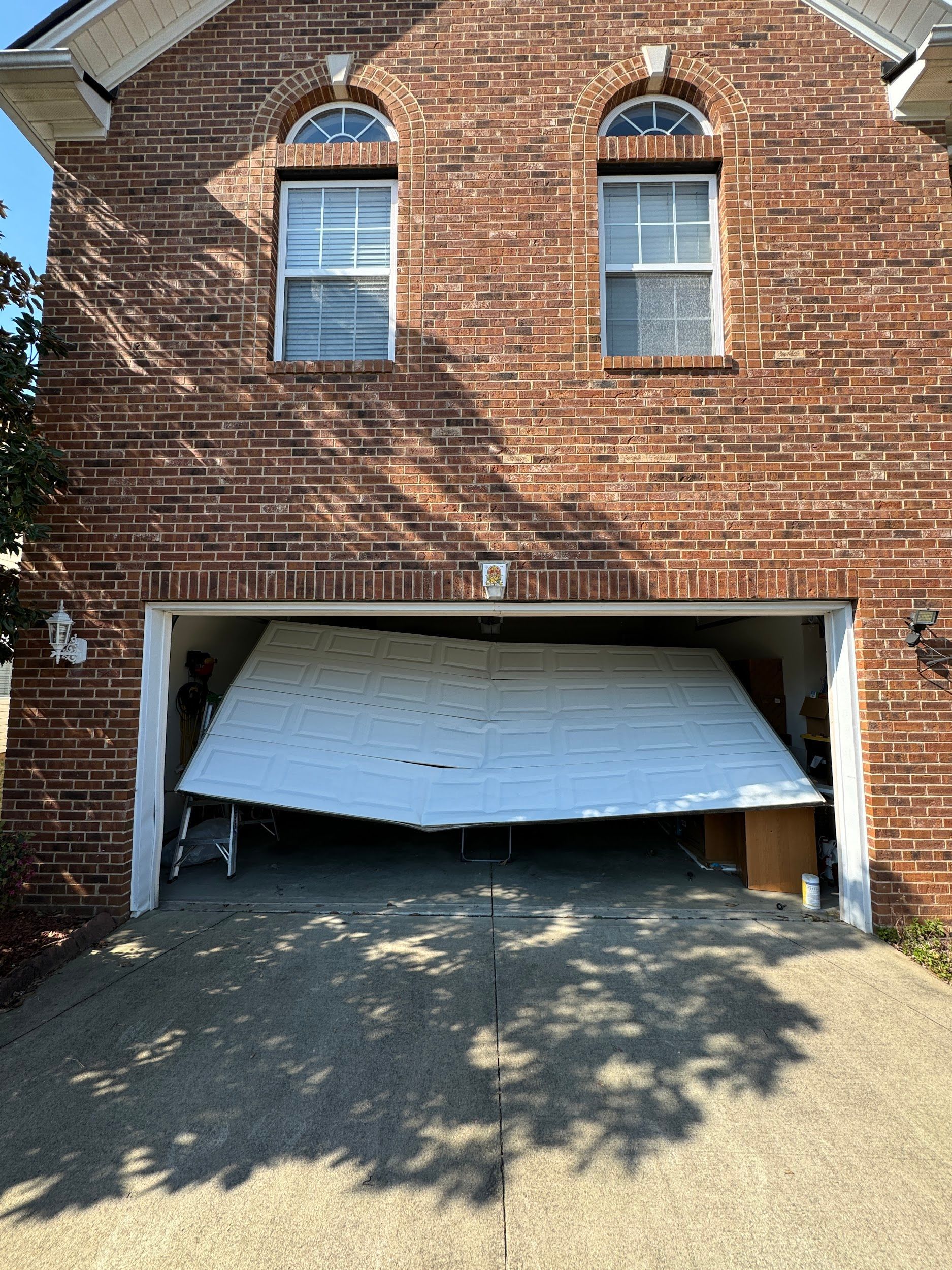 Garage door partially open, sagging. Red brick exterior. Shadowed driveway.