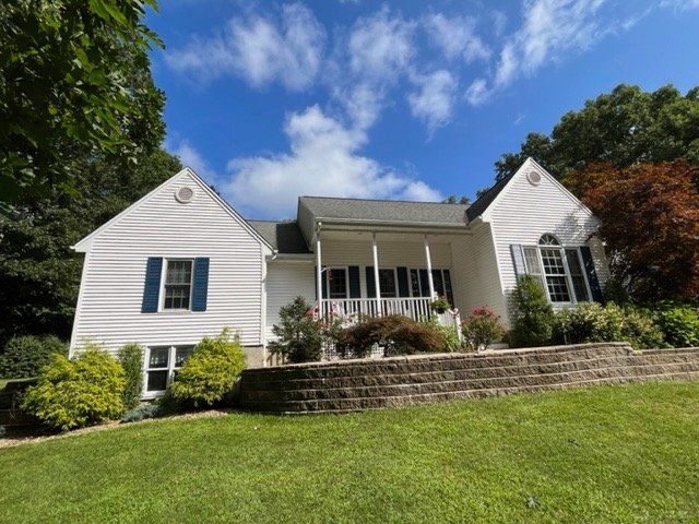 White house with gray roof and front porch on a sunny hillside, framed by trees and blue sky