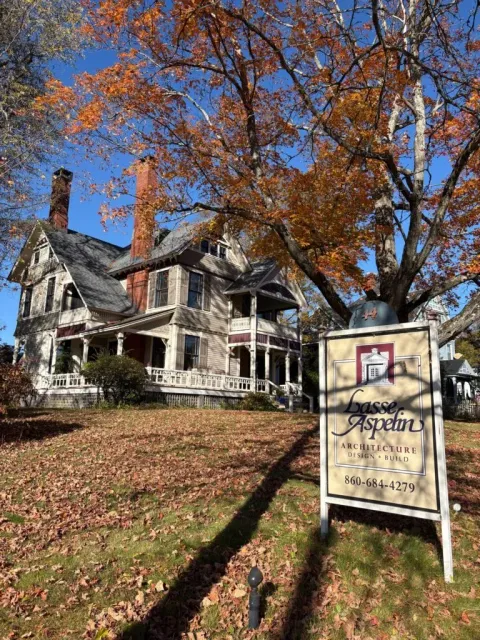 Historic house with white porch and autumn trees, plus a hanging sign in front on a sunny day