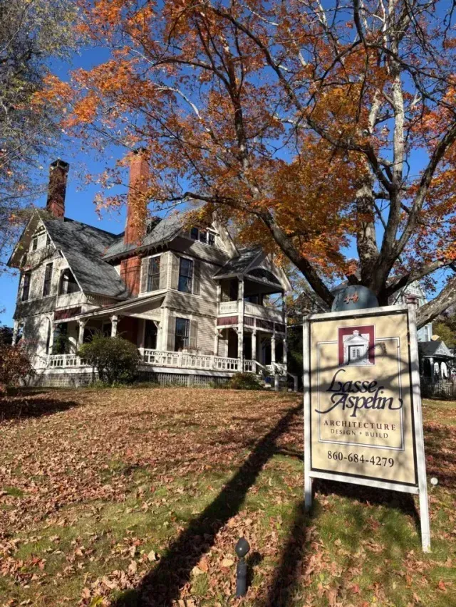 Historic house with white porch and autumn trees, plus a hanging sign in front on a sunny day