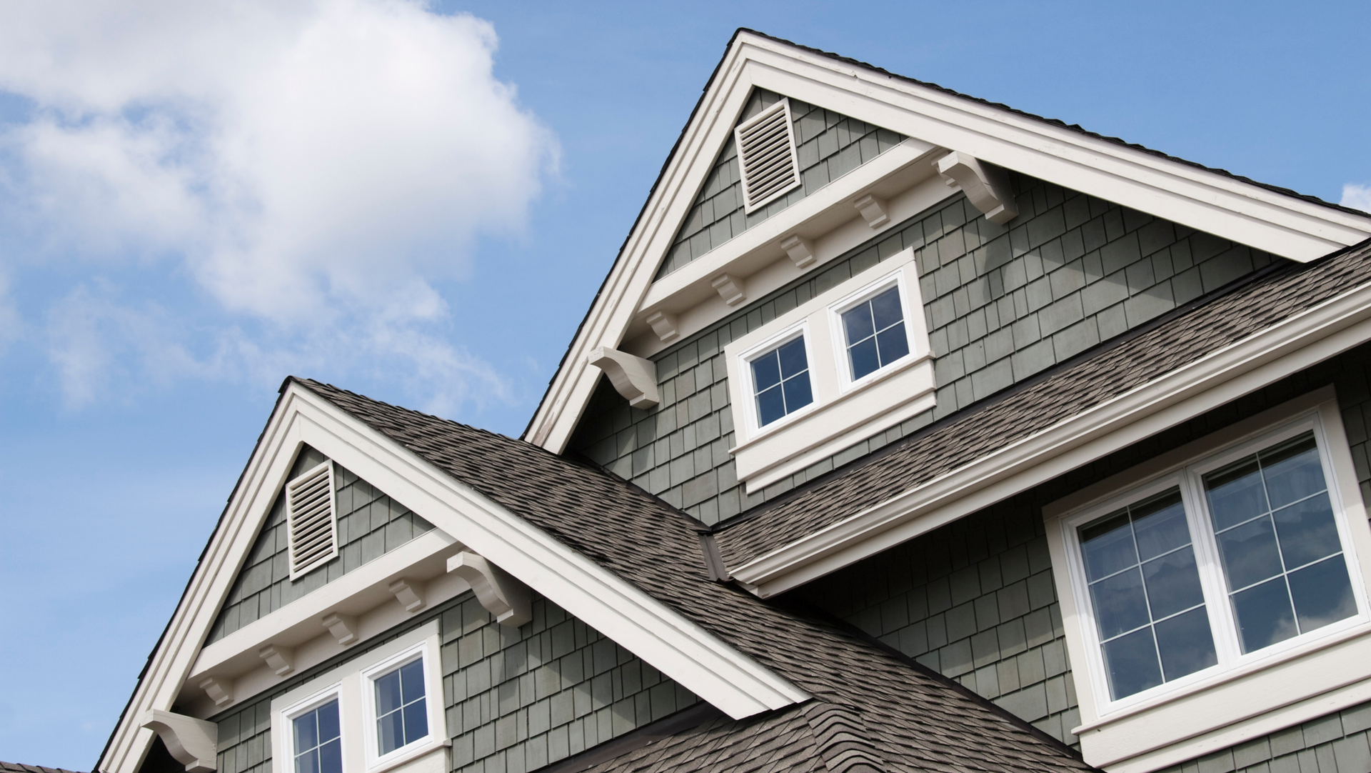 Two gray-shingled house rooftops with white trim and dormer windows against a blue sky