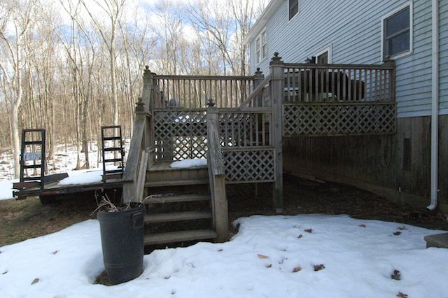 Snowy backyard deck with stairs, railing, and a house exterior beside bare winter trees