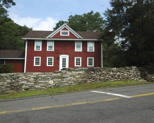 Red clapboard house with white trim behind a stone wall beside a road.