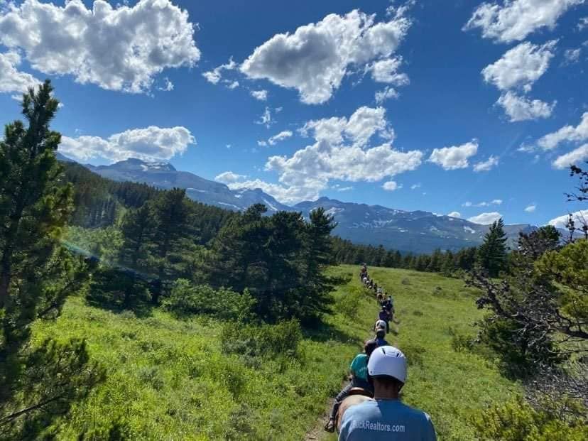 A Group of People Are Riding Horses Through a Grassy Field With Mountains in the Background