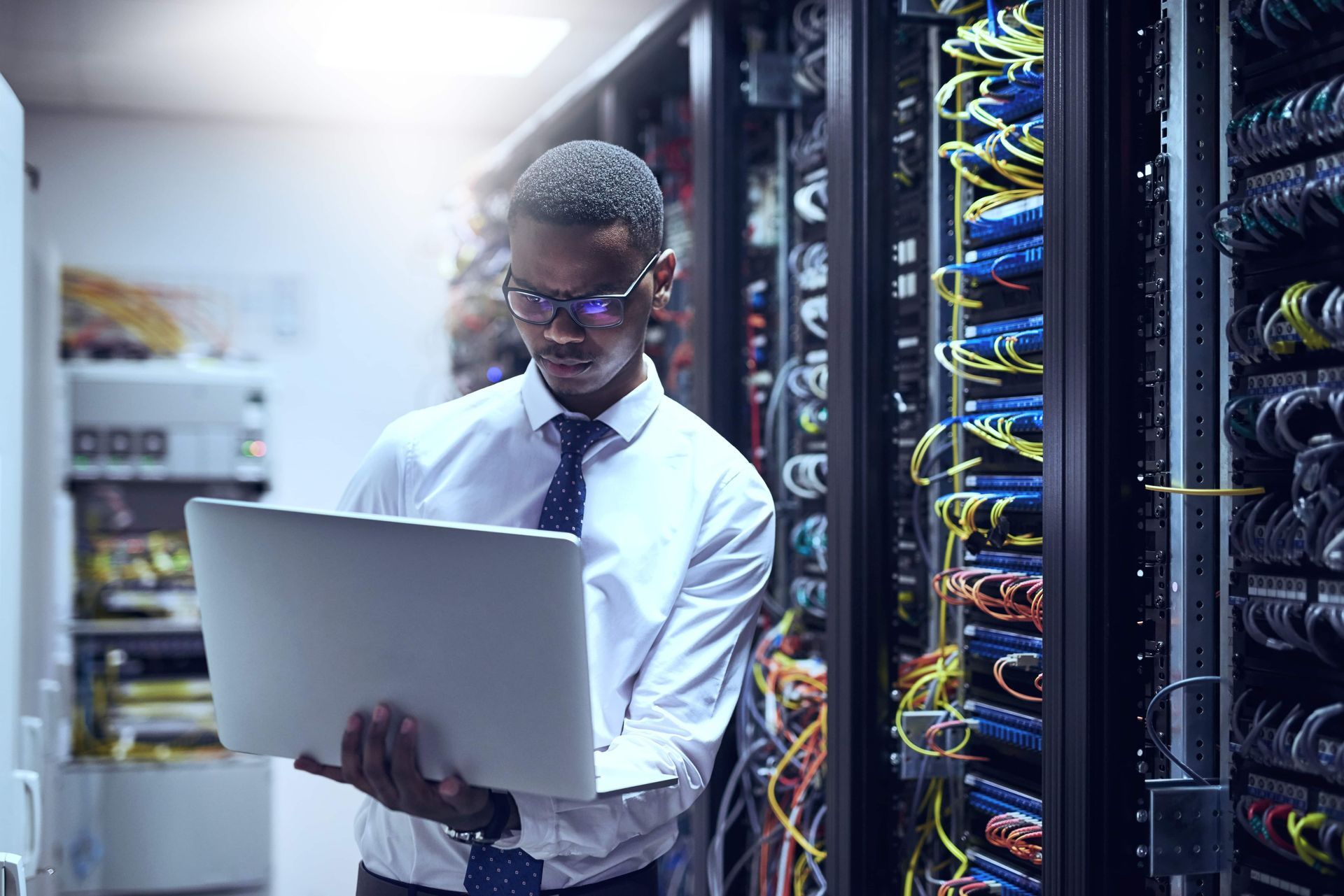 Man in server room, working on laptop, inspecting network cables.