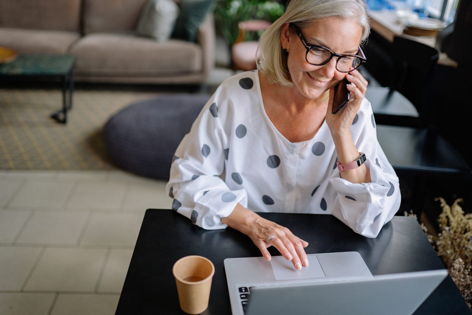 Woman with glasses on a laptop, smiling while on a phone call in a cafe.