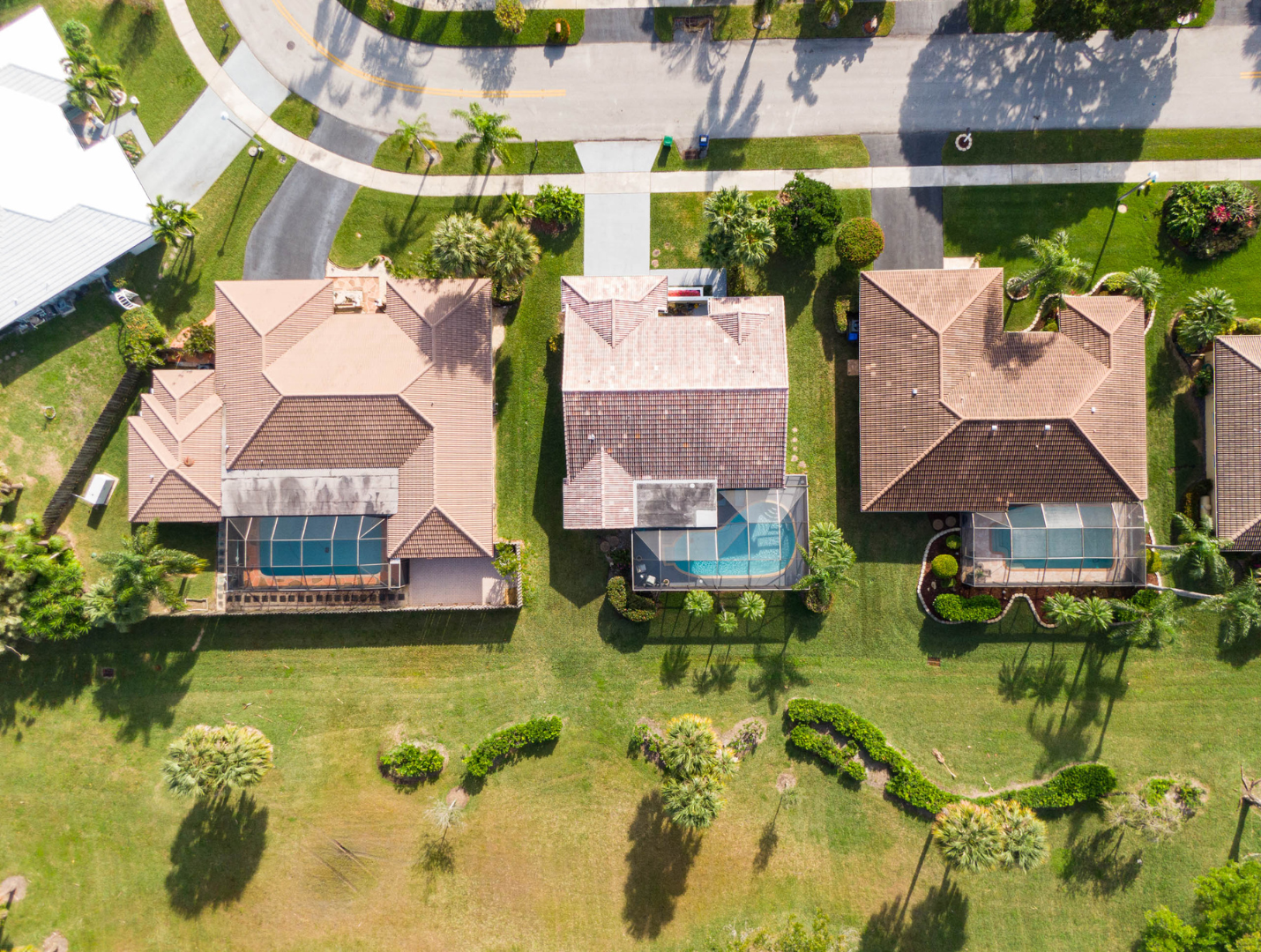 Aerial view of three houses with pools and tile roofs, surrounded by green lawns and palm trees.