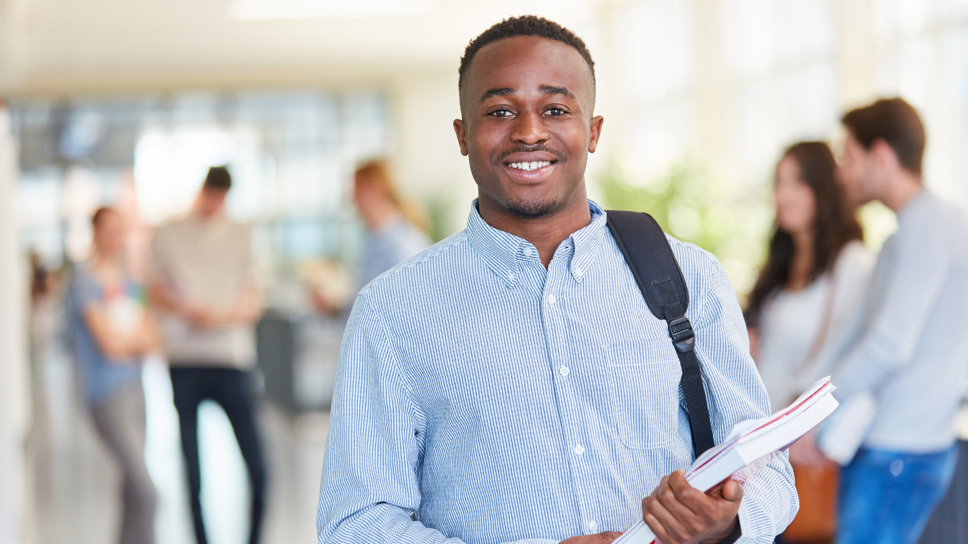 Smiling Black student with backpack and books in a hallway, other students in background.