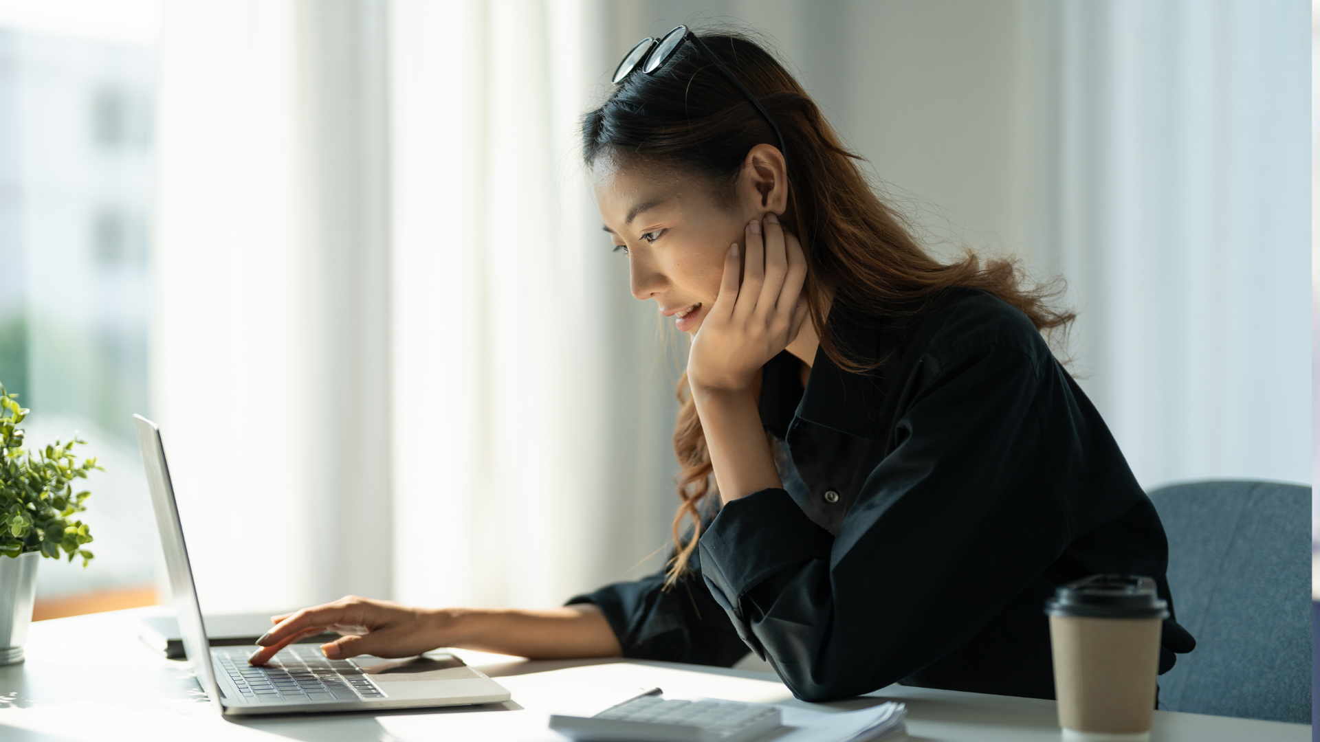 Woman at a desk with laptop, resting her head on hand, indoors, sunlight streaming through window.