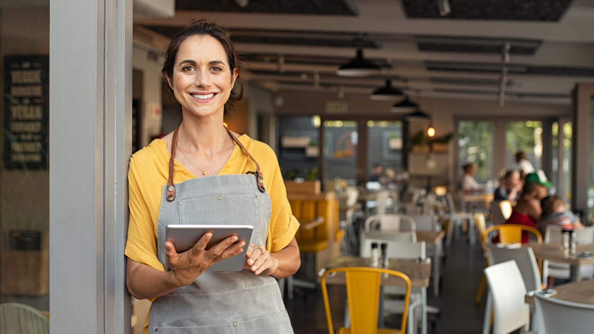 Woman in yellow shirt and gray apron, holding a tablet, smiling at cafe entrance.