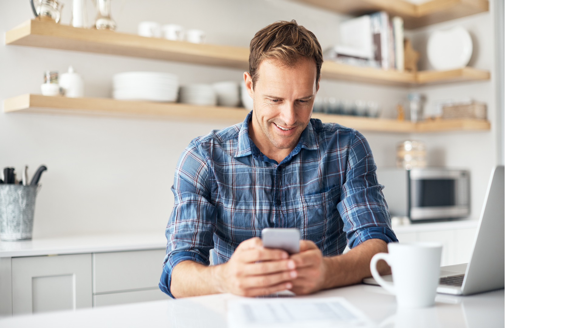 Man in blue plaid shirt smiles while looking at his phone in a kitchen with laptop and coffee cup.