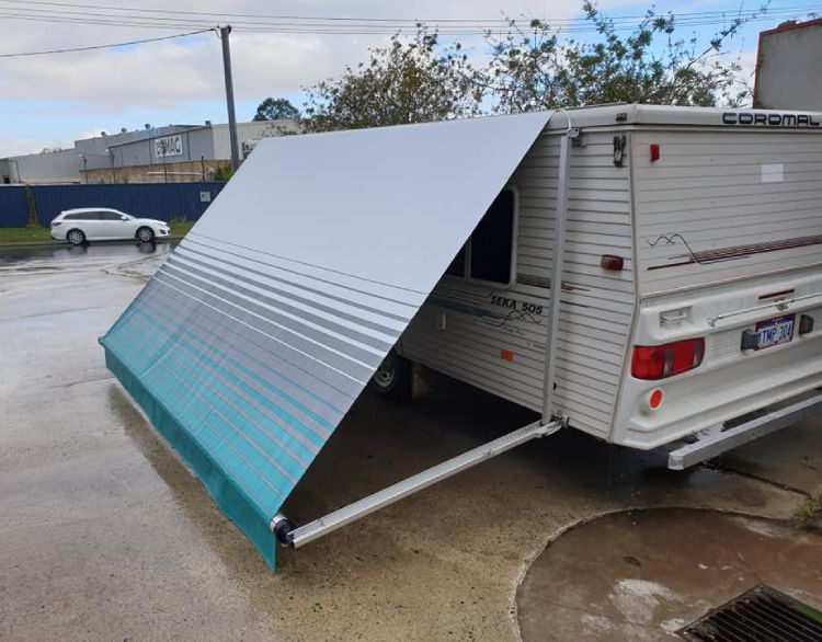 A caravan with a grey and blue striped awning extended, parked on a concrete surface with industrial buildings in the background.