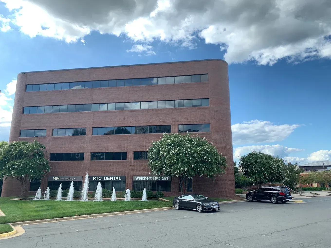 Brick office building with curved facade, fountain, trees, cars, and a cloudy blue sky.