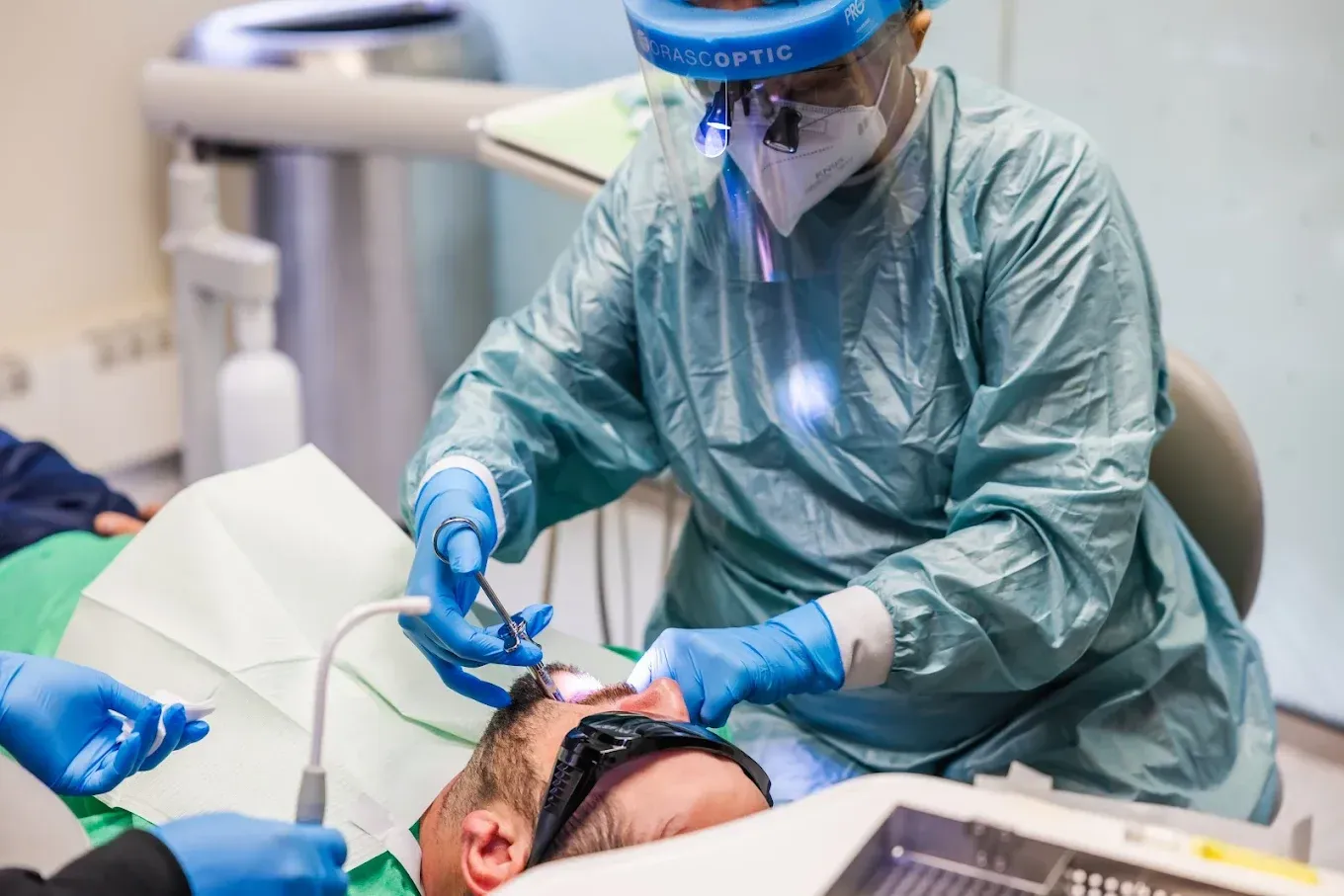 Dentist in protective gear performing a procedure on a patient in a dental chair.