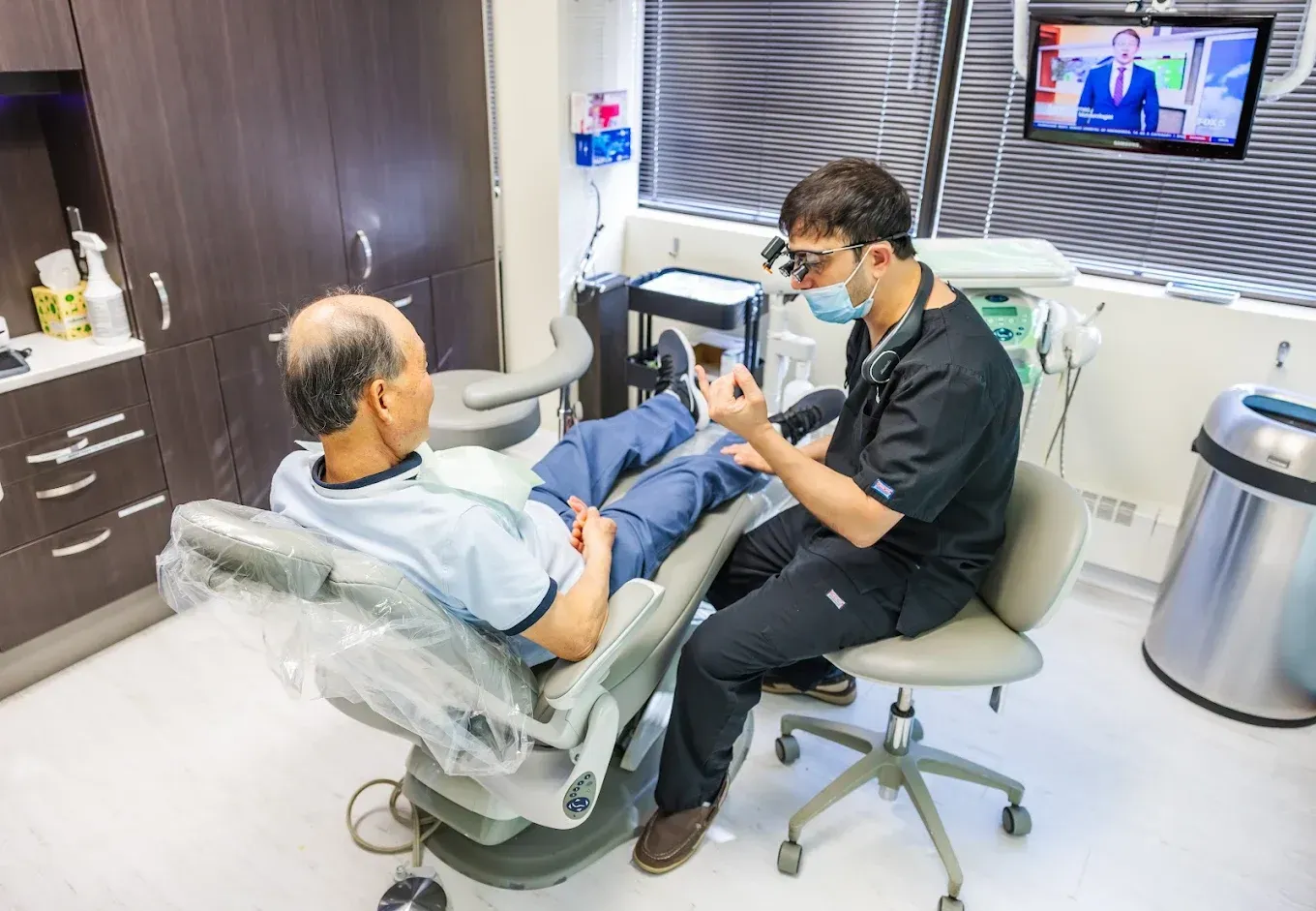 Dentist showing something to a patient seated in a dental chair in a clinic; a TV is on in the background.