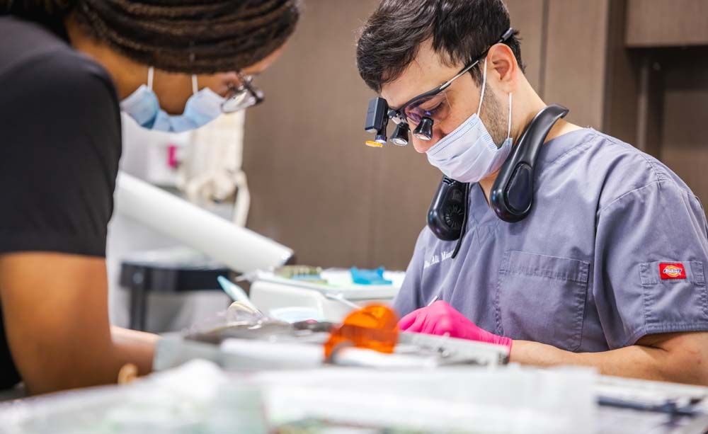 Two medical professionals in scrubs, working on a dental procedure. One wears glasses and a magnifying device.