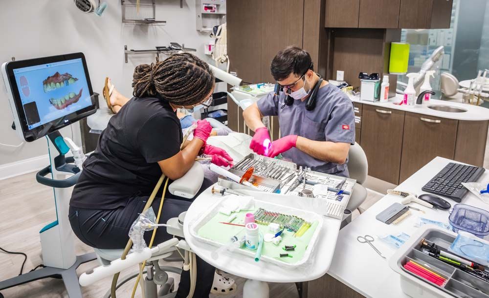 Dentist and assistant working on a patient in a dental office. Both wear masks, gloves, and scrubs.
