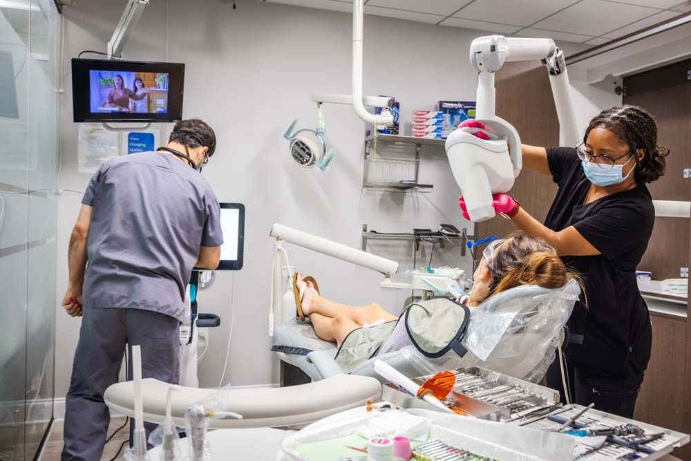 Dentist and assistant preparing dental x-ray for patient in a clinic room; light blue and white setting.
