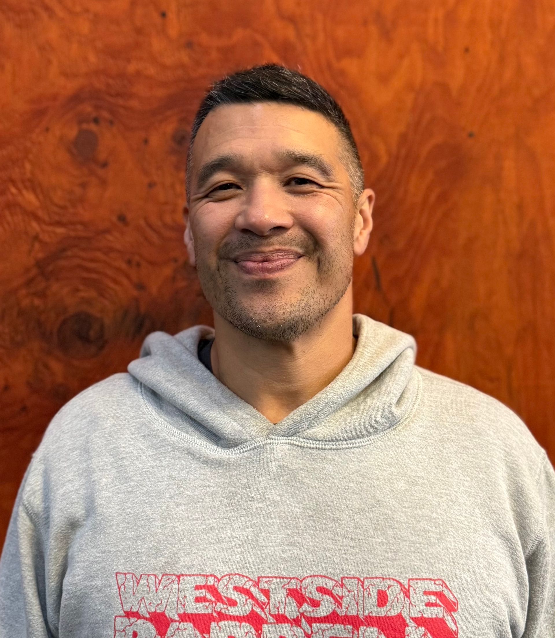 Man in grey hoodie smiles, with a wooden background.