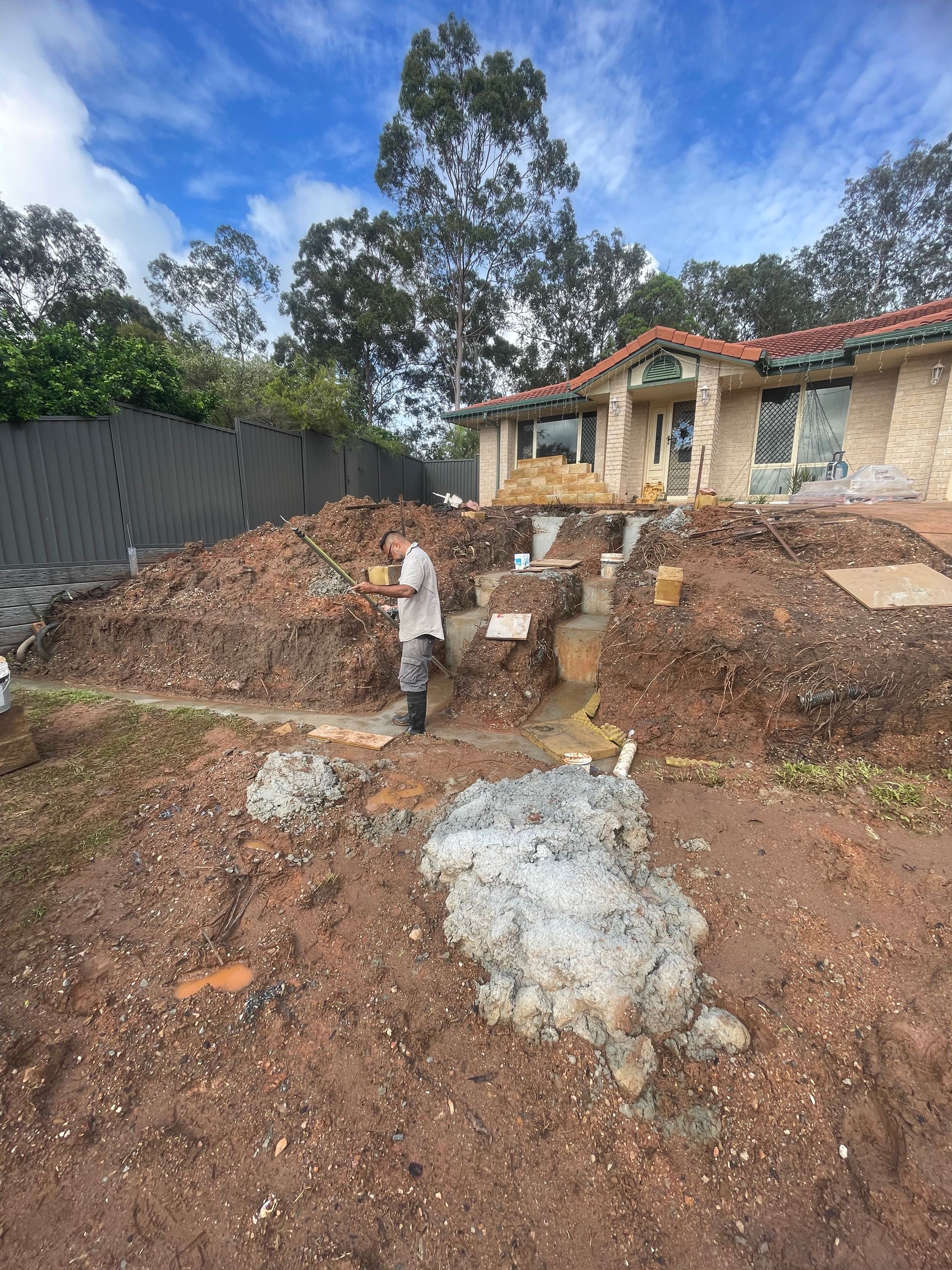 Construction site. A man standing on dirt, near a house with exposed foundations. Overcast sky.