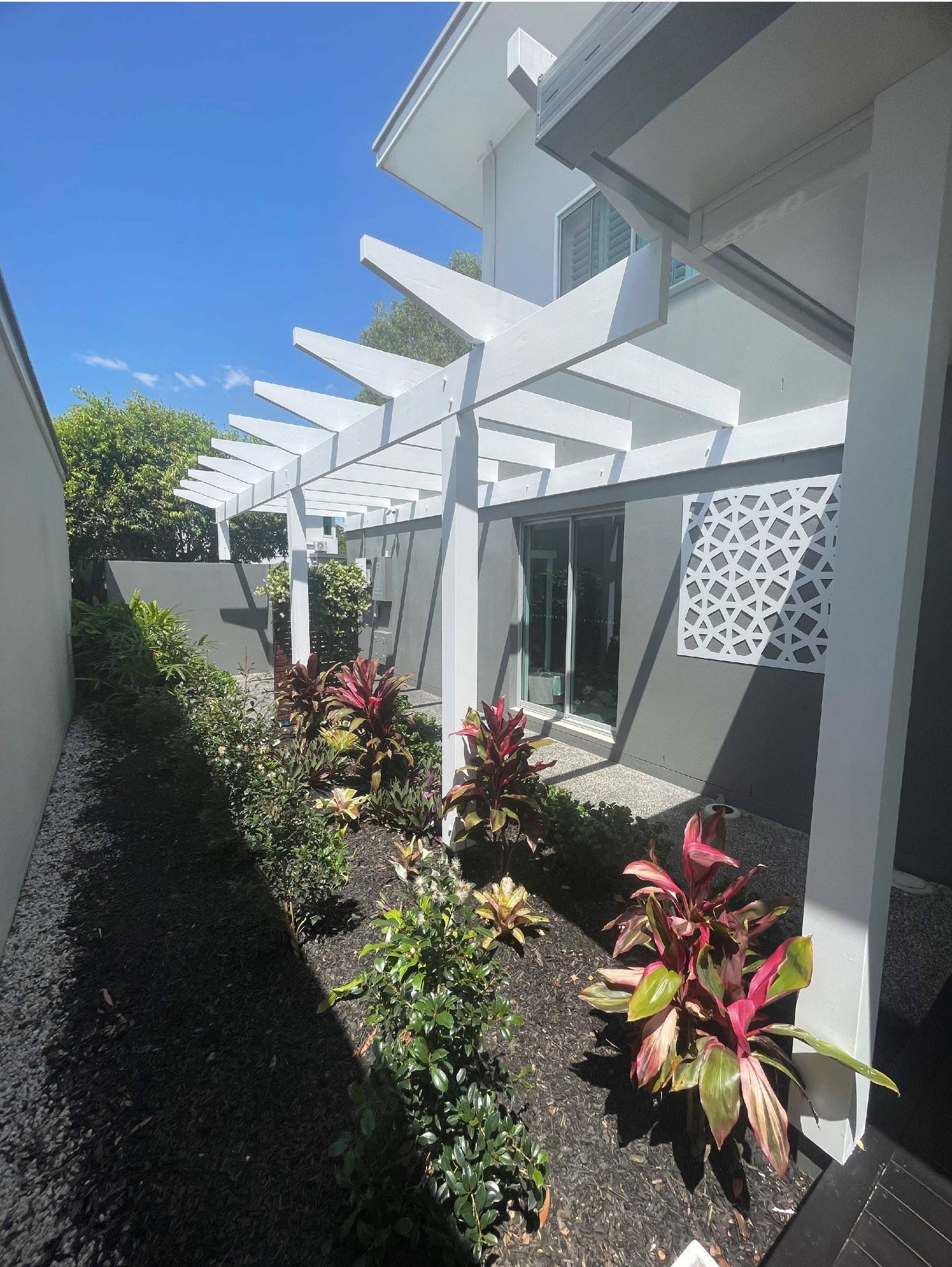 White pergola over a narrow garden with lush red and green plants; sunny day.
