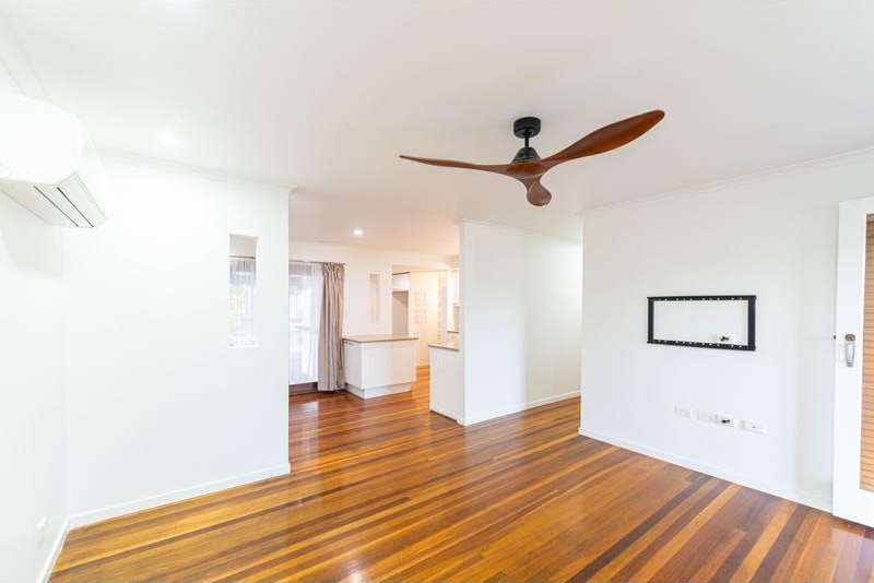 Bright room with wooden floor, white walls, and a ceiling fan, leading to a kitchen area.