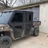 Black off-road utility vehicle parked beside a beige garage, with oversized tires and camo-pattern accents.