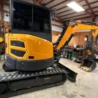 Yellow compact excavator with black tracks parked inside a workshop, side bucket visible