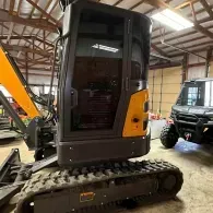 Compact excavator with black cab and tracked base inside a warehouse, beside a utility vehicle