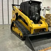 Yellow compact tracked loader with front bucket indoors in a warehouse.