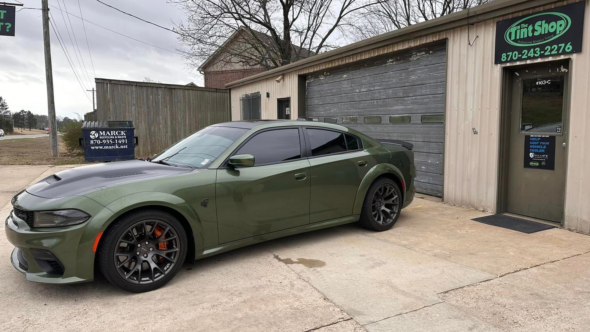 Olive-green Dodge Challenger parked outside a tire shop with a sign and garage door nearby