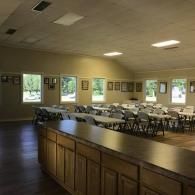 Empty banquet hall with rows of chairs, long tables, and a wooden serving counter by large windows.