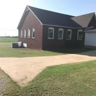 Brick building with arched windows and a sloped roof beside a grassy yard and paved driveway
