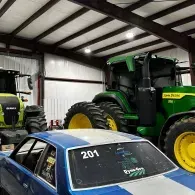 Blue race car in a garage beside a green tractor and yellow tractor tires