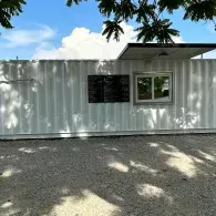 White shipping container building with a window and sign, seen under tree shade on a gravel lot.