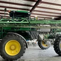 Large green agricultural sprayer with yellow wheels inside a barn-like shed